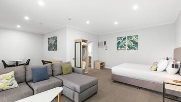 A modern hotel room with a grey sectional sofa, a single bed, a small table, and a mirror.