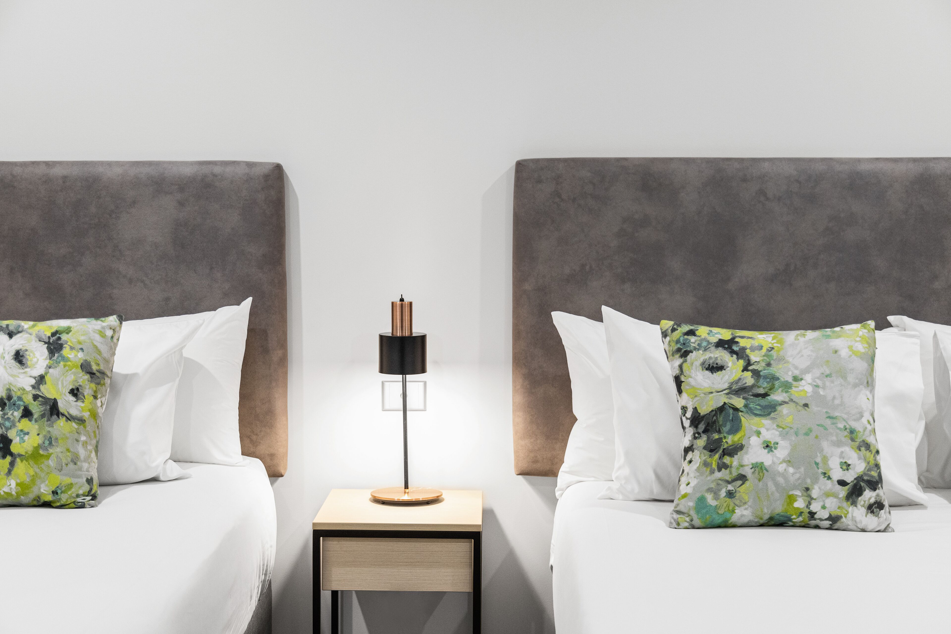 Hotellihuone, jossa on kaksi sänkyä, yöpöytä lampun kanssa ja kukkakuviolliset tyynyt.