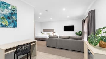 A modern hotel room with a desk, a grey sofa, a bed, and a large window with curtains.