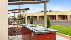 An outdoor kitchen area with a sink and a fire extinguisher, surrounded by a covered patio with a view of a parking lot and a building in the background. - Presidential Motel (Mount Gambier)