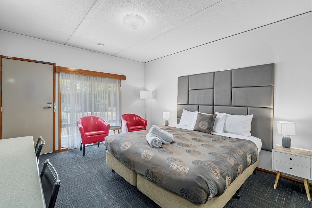 Una habitación de hotel con una cama grande, un escritorio, una silla, una butaca roja, una mesita de noche y una lámpara.
