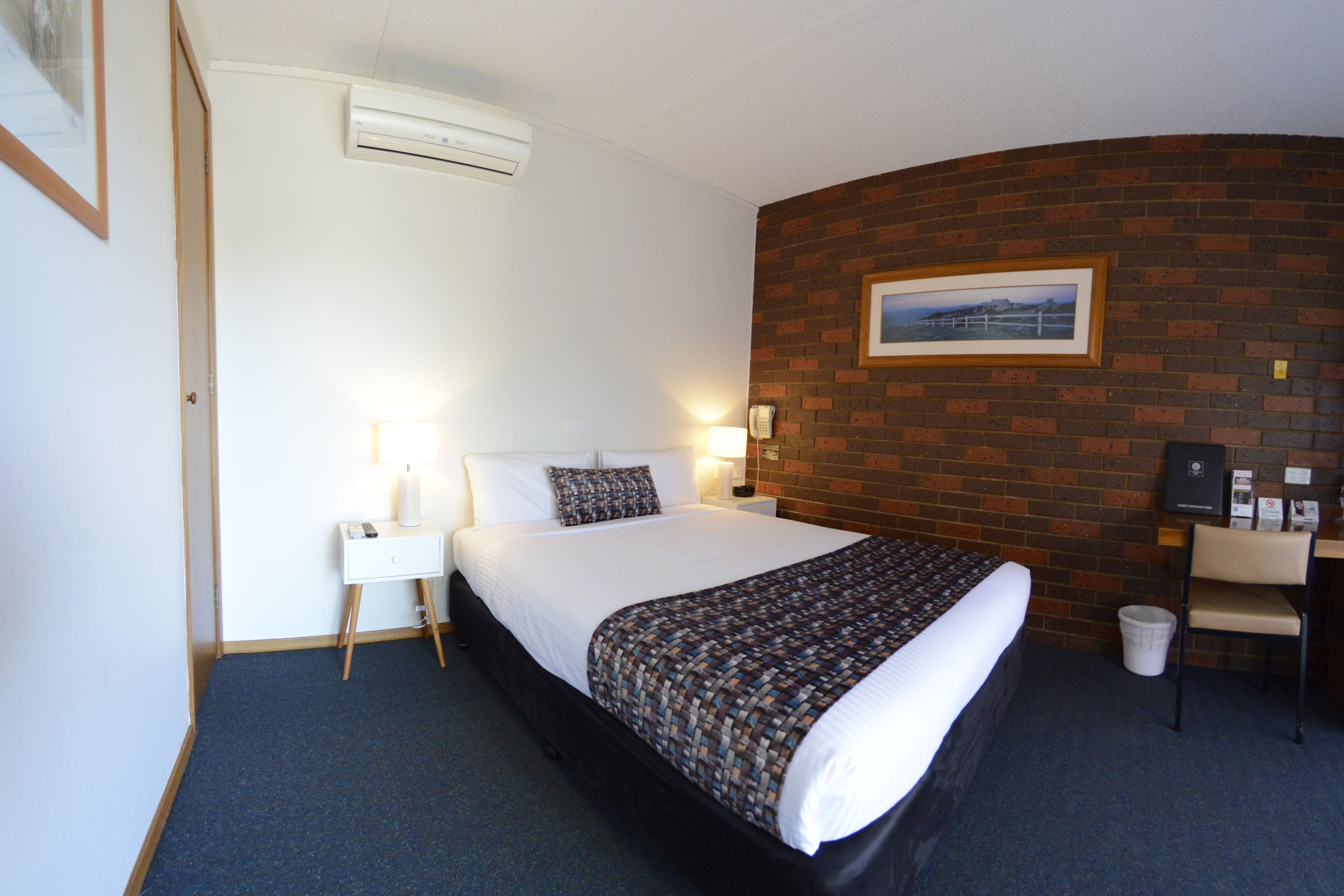 Quarto de hotel com cama, mesa de cabeceira, abajur e escrivaninha com cadeira.