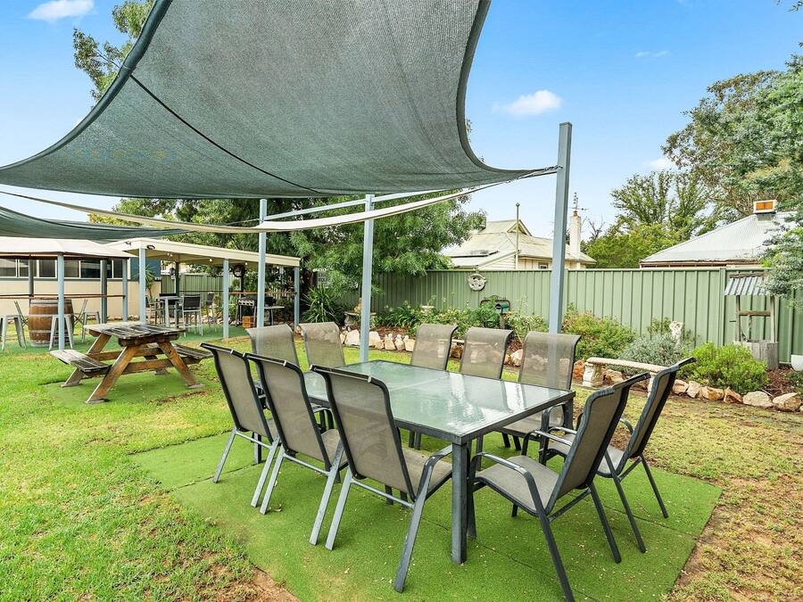 An outdoor patio with a glass table and chairs, surrounded by a green lawn and a shaded area.