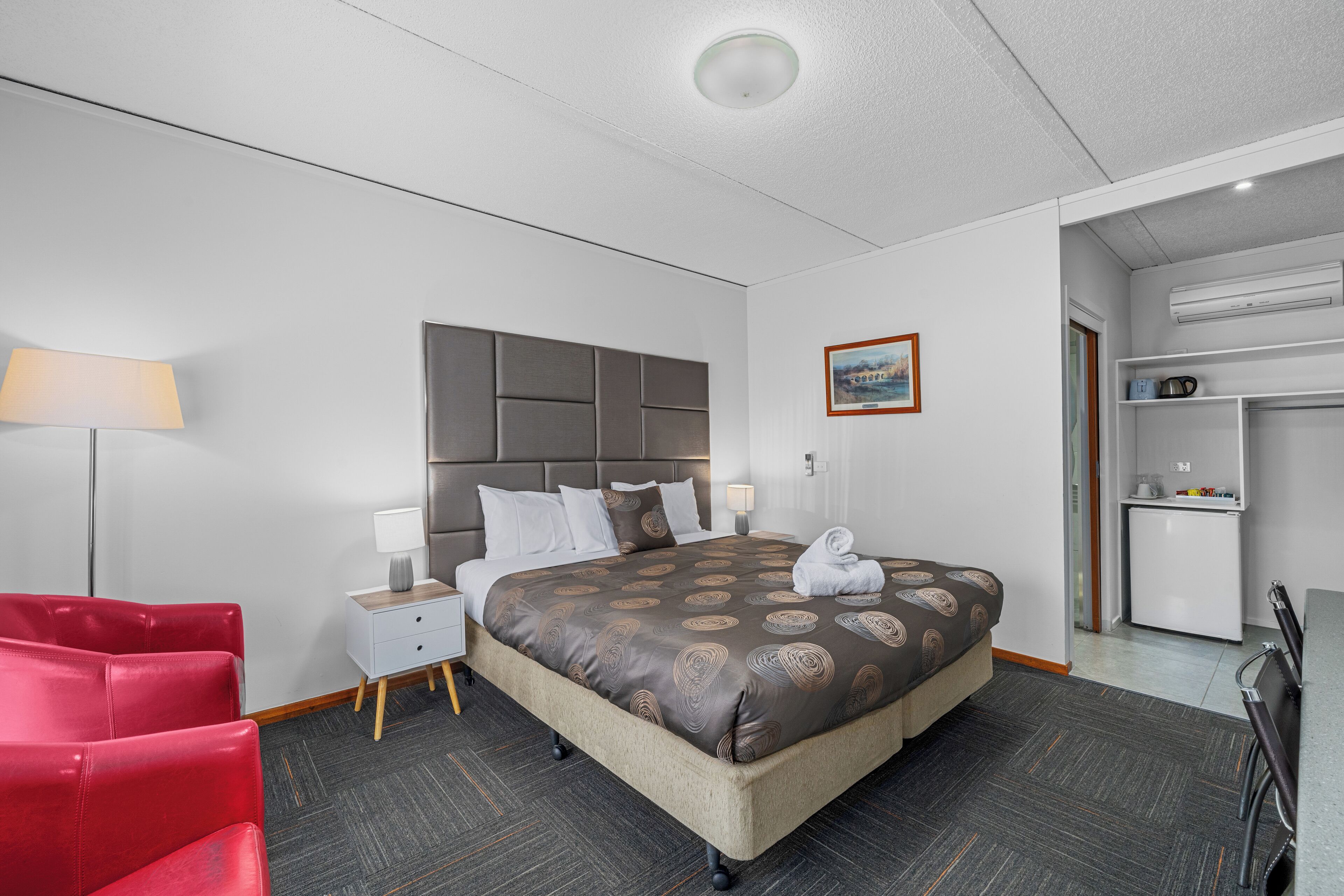 A hotel room with a large bed, a red chair, a small desk, and a kitchenette.