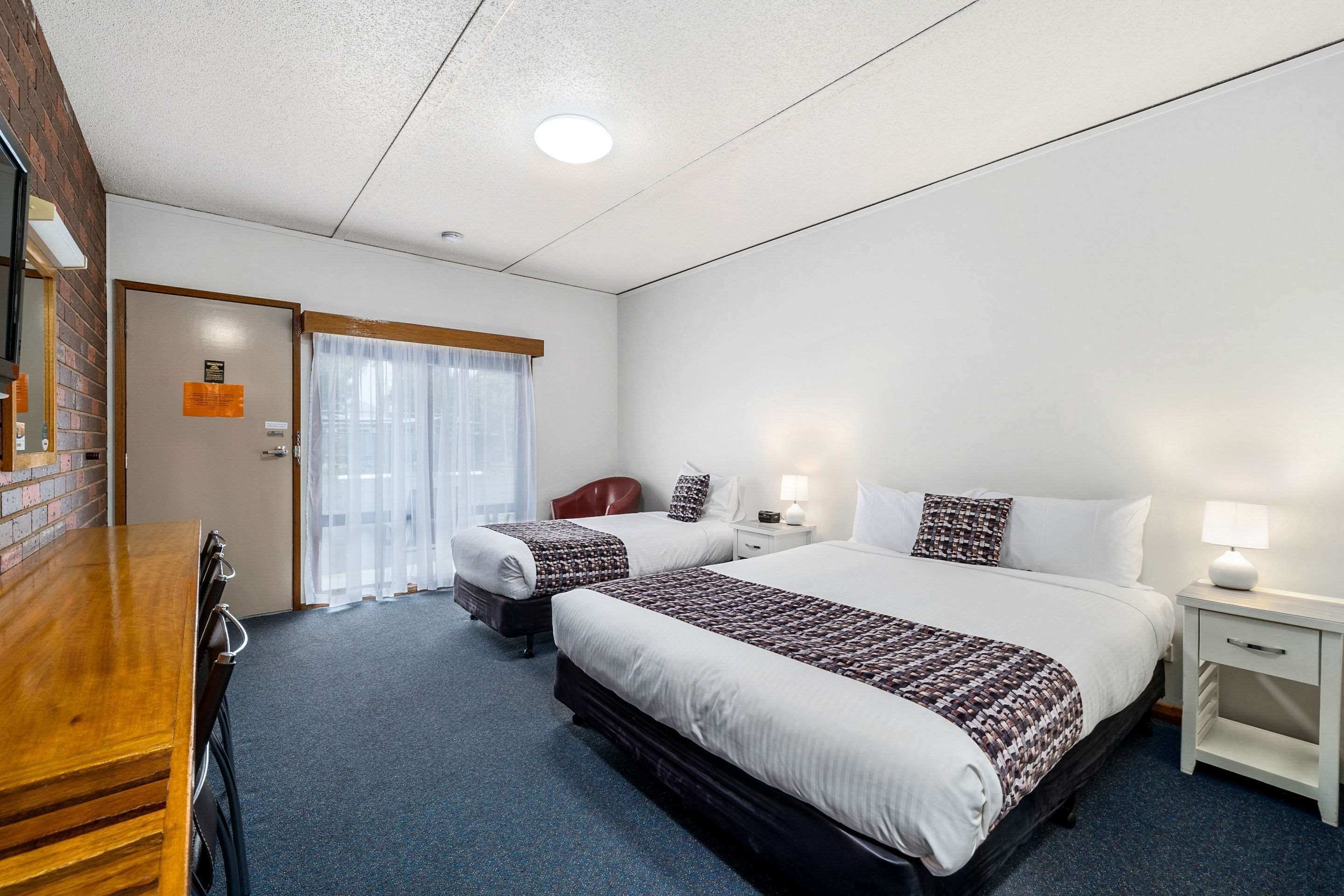 Une chambre d’hôtel avec deux lits, un bureau en bois, une table de chevet blanche et une fenêtre avec des rideaux légers.