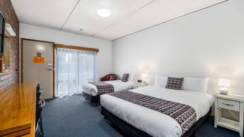 A hotel room with two beds, a wooden desk, a white nightstand, and a window with sheer curtains.