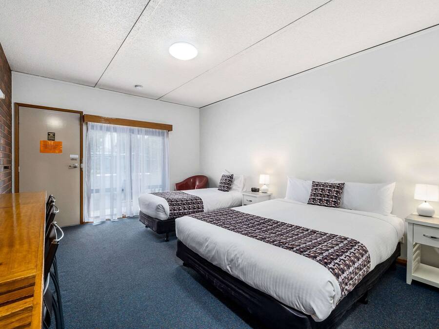 A hotel room with two beds, a wooden desk, a white nightstand, and a window with sheer curtains.