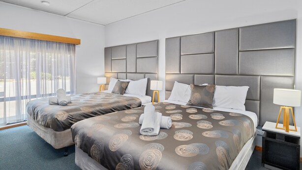 Ein Hotelzimmer mit zwei Betten, einem großen Fenster mit Vorhängen, einem Kopfteil mit Strukturmuster und Nachttischlampen.