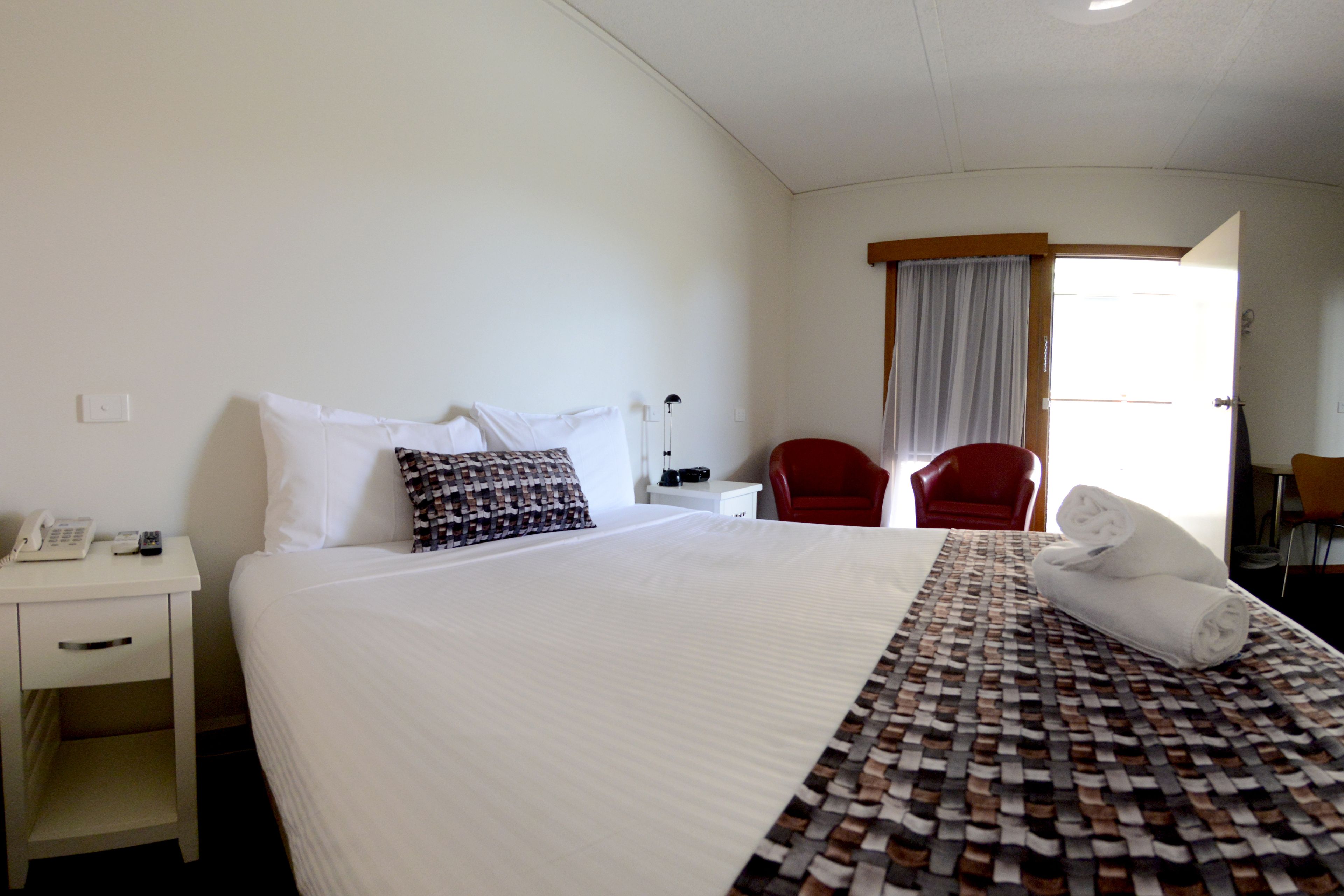 Quarto de hotel com uma cama grande, uma mesa de cabeceira com telefone, uma cadeira vermelha e uma janela com cortinas.