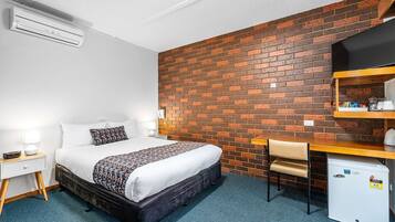 A hotel room with a brick wall, a bed with a patterned blanket, a desk with a chair, a small refrigerator, and a nightstand with a lamp.