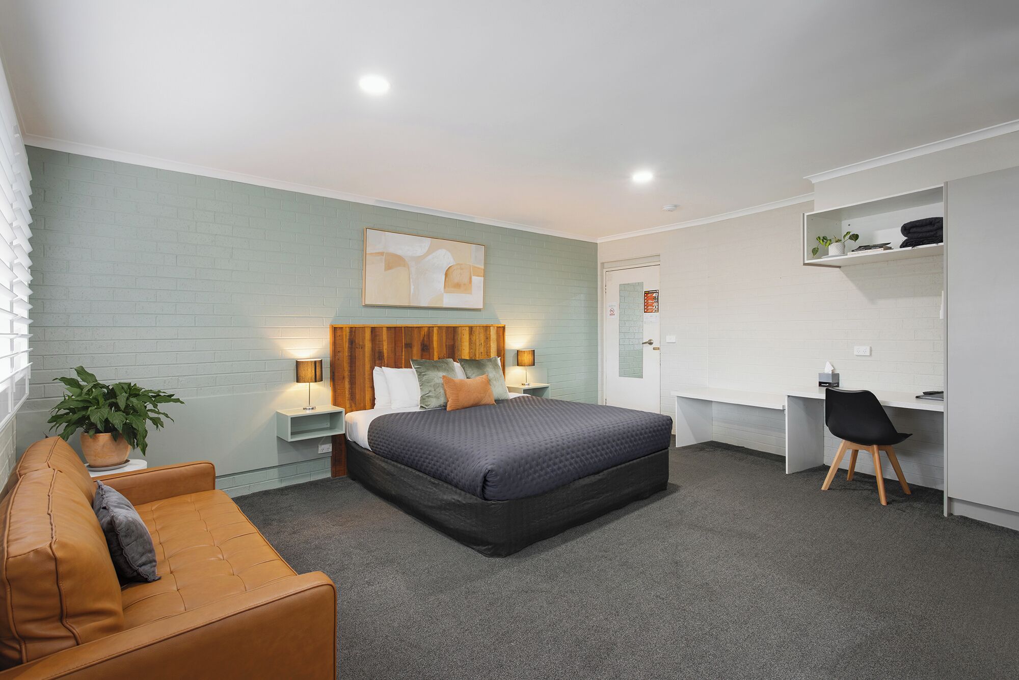 A modern hotel room with a bed, a desk, a chair, a sofa, and a potted plant.