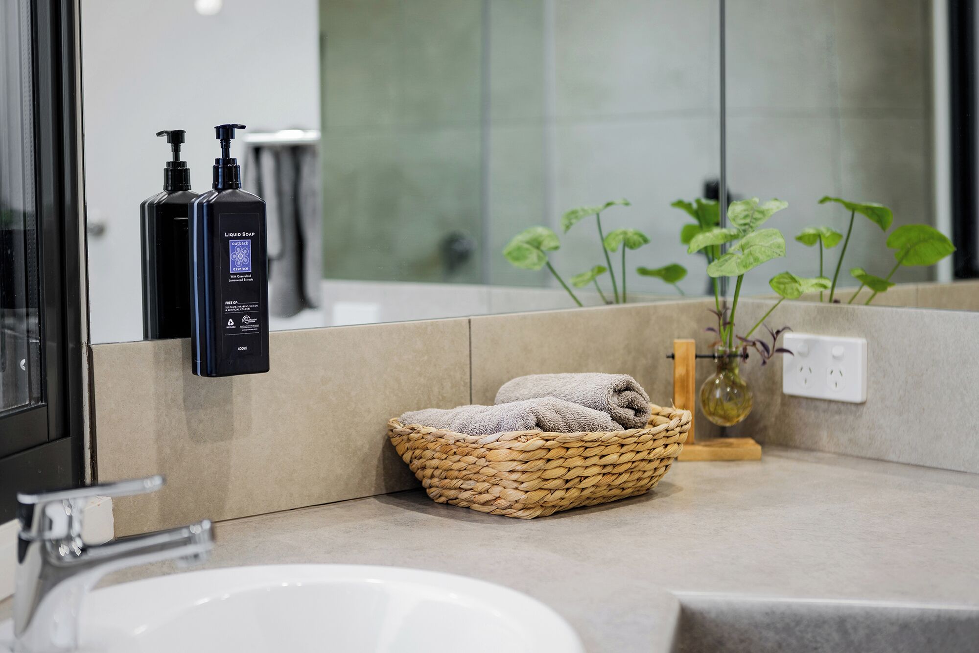 Un baño con lavamanos blanco, un espejo, un dispensador de jabón negro y una cesta de mimbre con toallas.