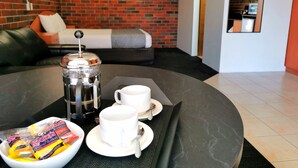 Una preparación de café con una prensa francesa, dos tazas y un cuenco de galletas sobre una mesa en una habitación con pared de ladrillo y un sofá al fondo.