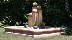 Una fuente de piedra con elementos esféricos y plantas en macetas en un jardín ajardinado.