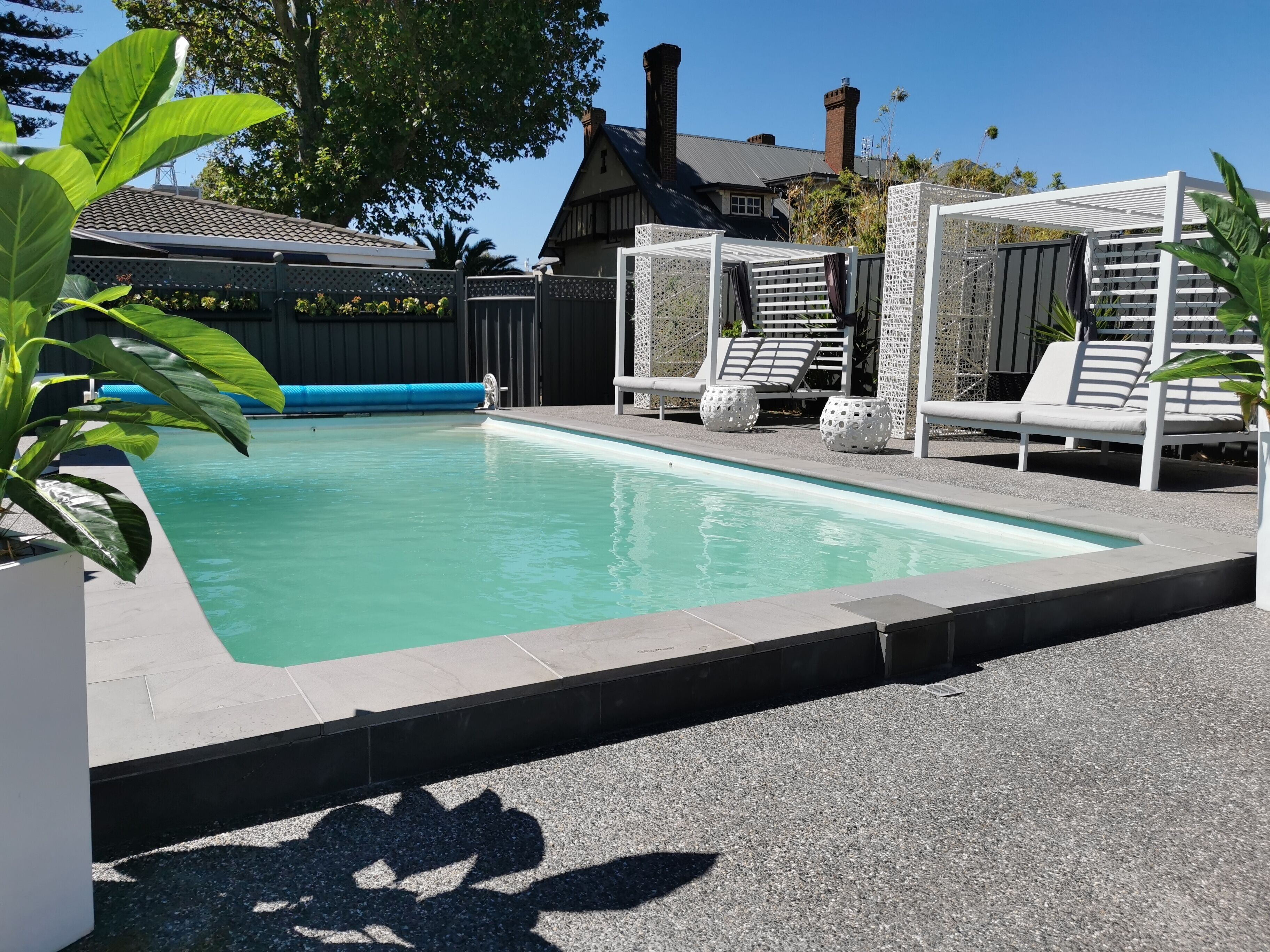 A rectangular swimming pool with a tiled edge, surrounded by a paved deck. There are white lounge chairs and a covered seating area with a white lattice structure.