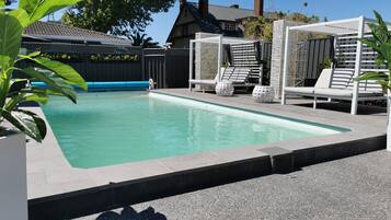 A rectangular swimming pool with a tiled edge, surrounded by a paved deck. There are white lounge chairs and a covered seating area with a white lattice structure.