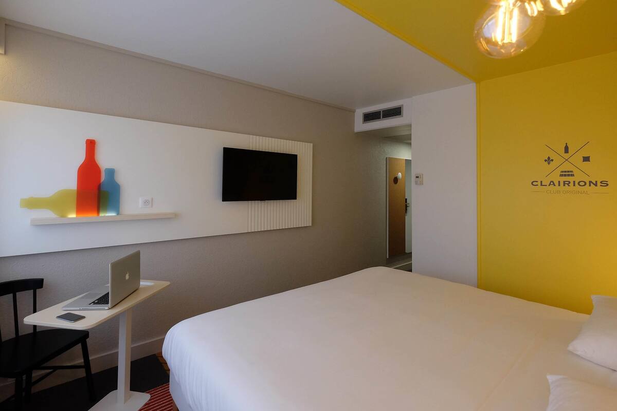 Ein Hotelzimmer mit Bett, Schreibtisch mit Laptop, Fernseher an der Wand und einem einzigartigen Kunstwerk.