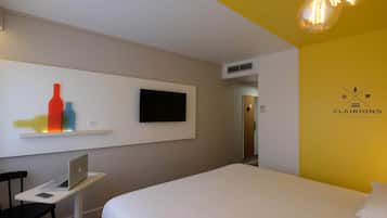 A hotel room with a bed, a desk with a laptop, a TV on the wall, and a unique wall art piece.