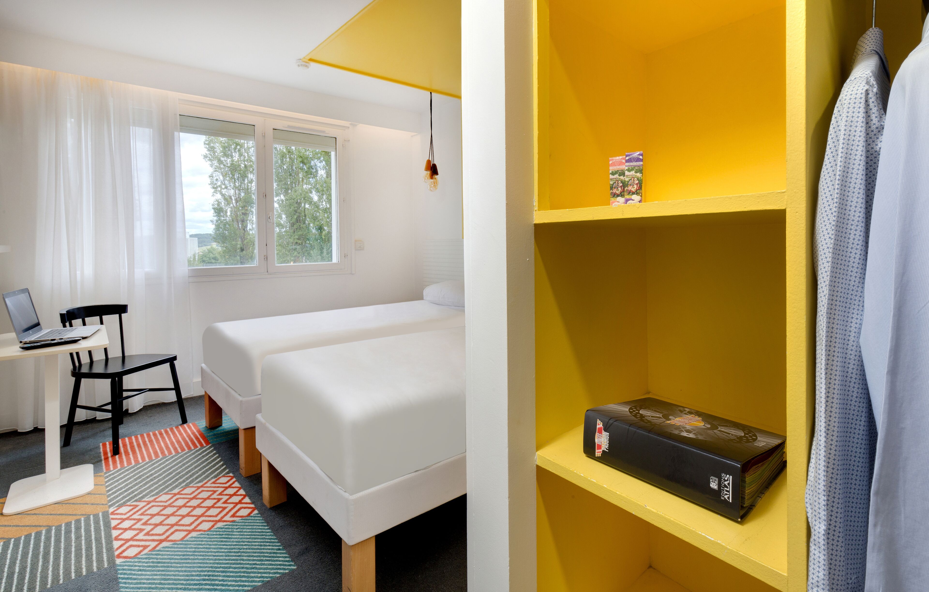 A hotel room with two beds, a desk with a laptop, a chair, and a bookshelf with a book.