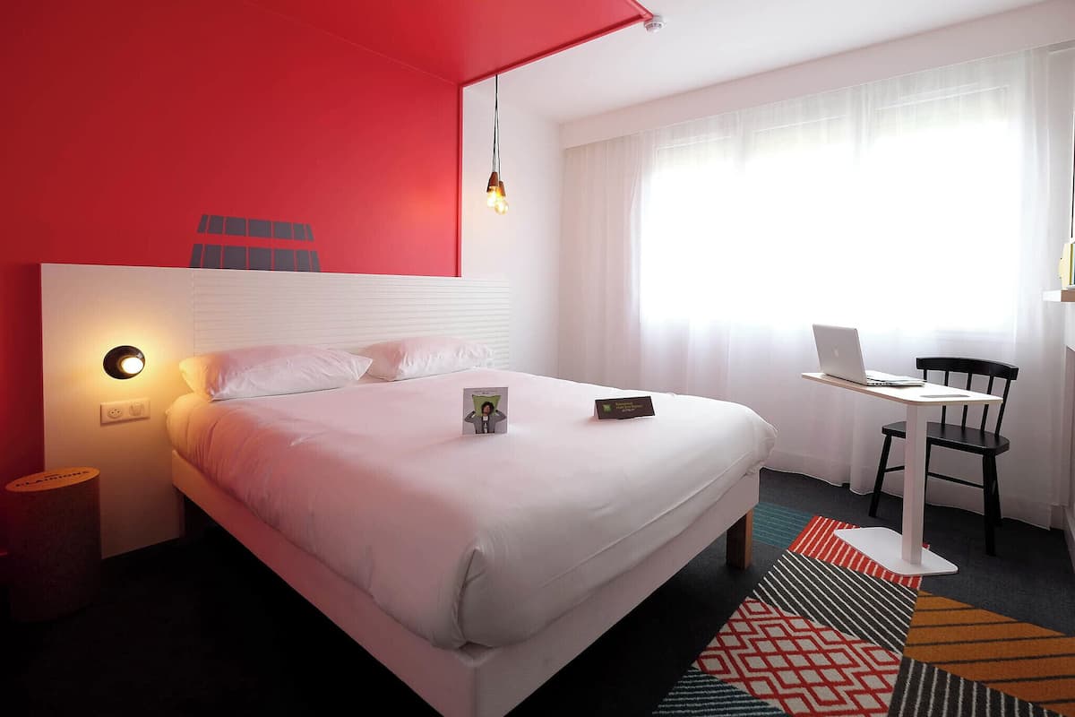 Ein Hotelzimmer mit Bett, Schreibtisch mit Laptop, Stuhl, Nachttisch mit Lampe und Fenster mit durchsichtigen Vorhängen.