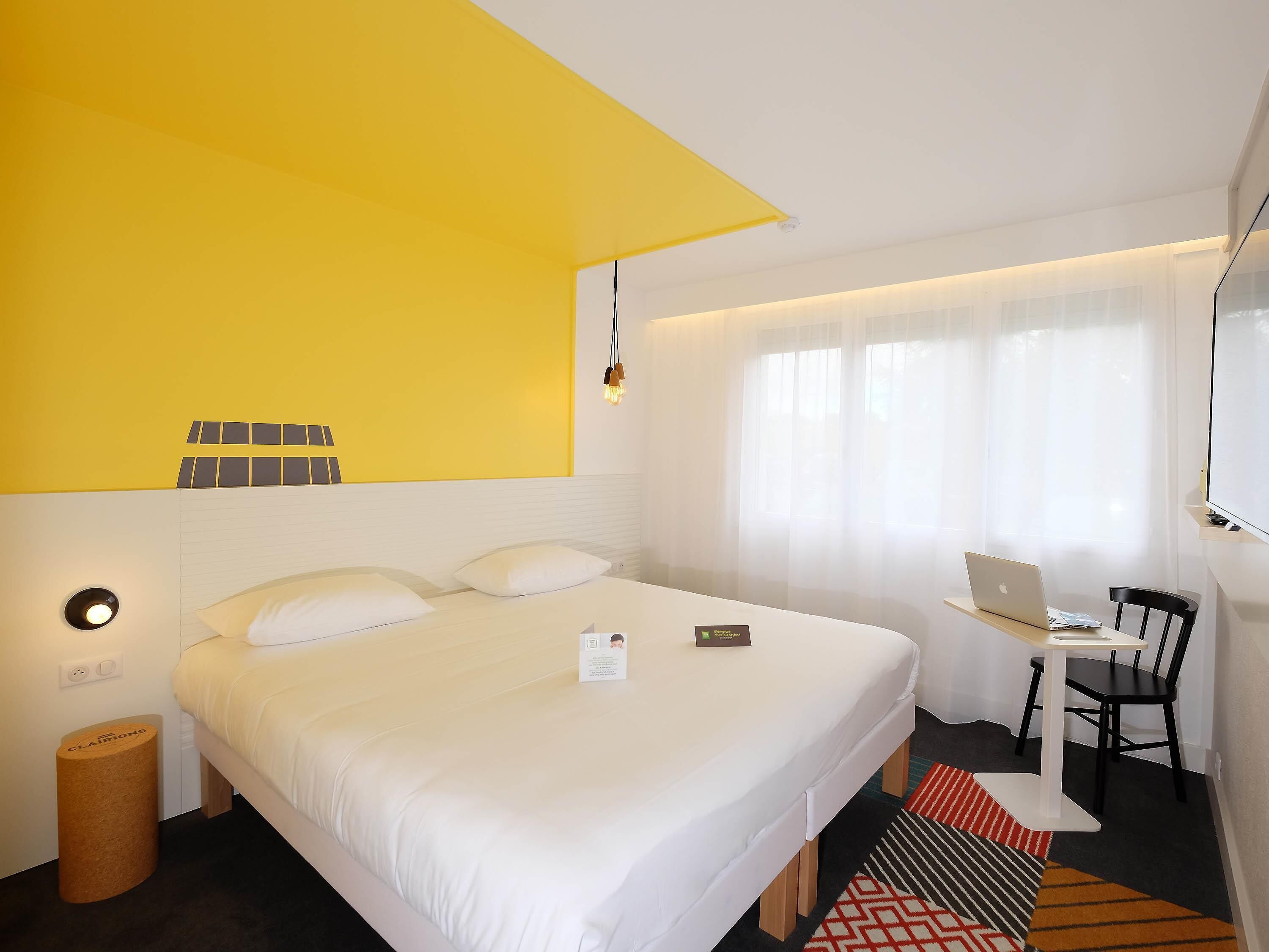 Habitación de hotel con cama, escritorio con una computadora portátil, silla, un cesto de basura pequeño y una ventana con cortinas.