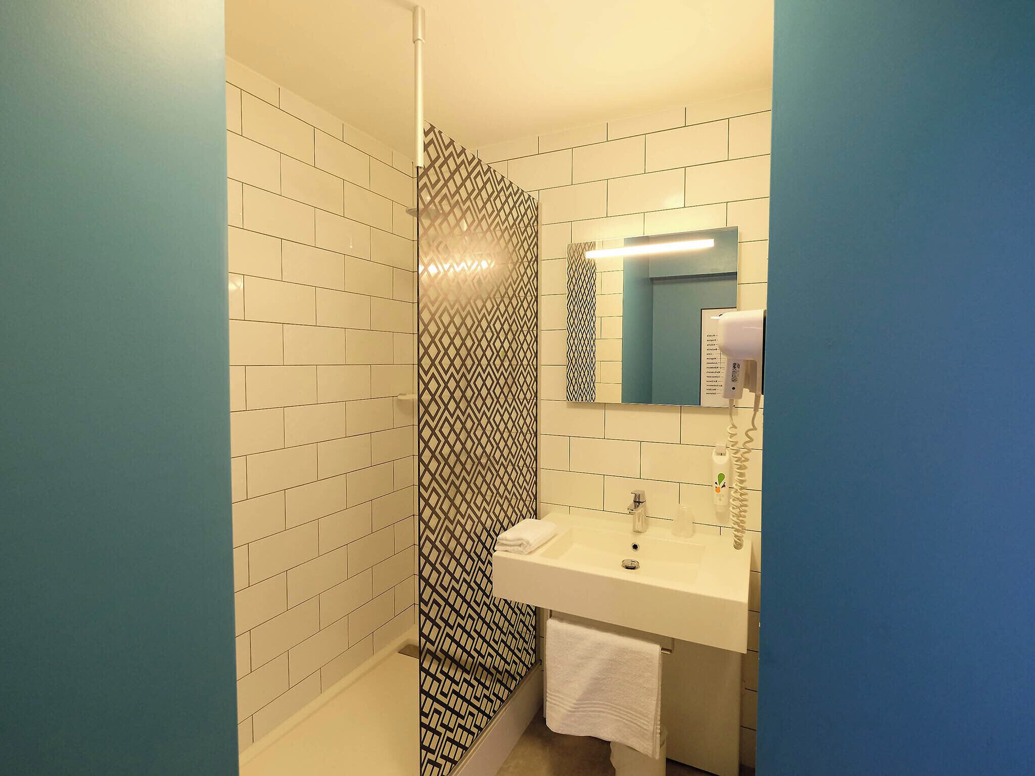 A bathroom with white tiled walls, a patterned shower curtain, a sink with a mirror, and a hairdryer.