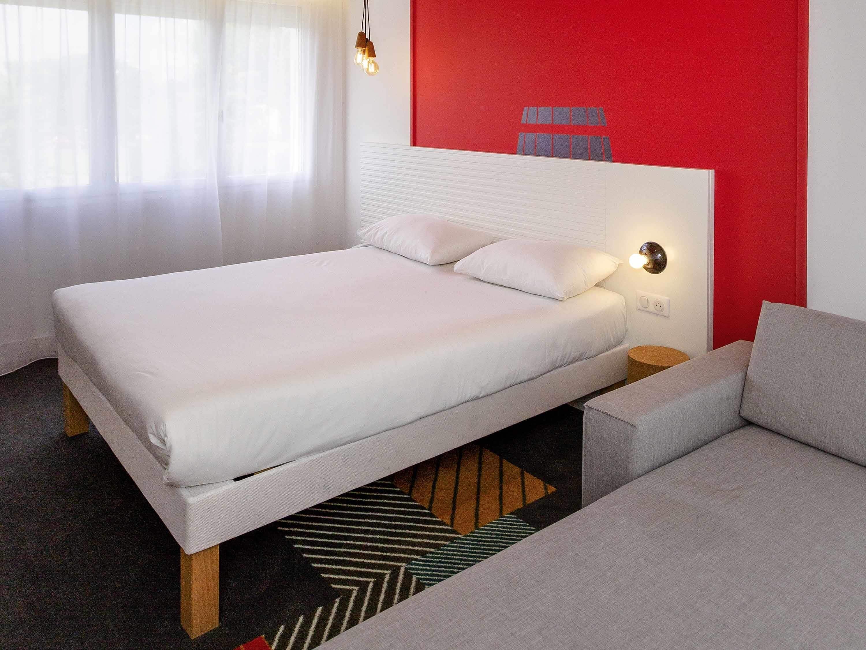 A modern hotel room with a large bed, a bedside table, a lamp, and a red accent wall.