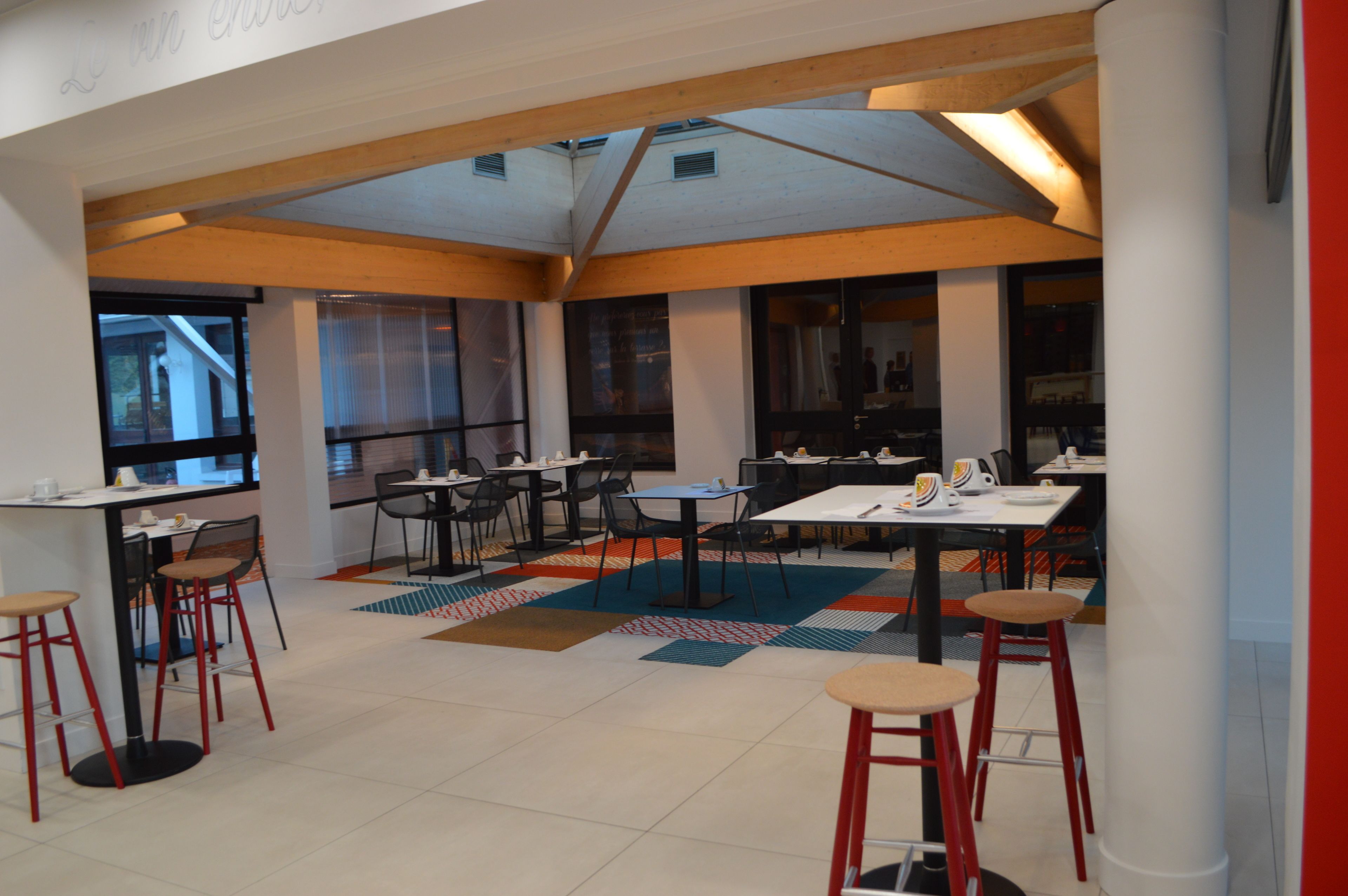 A modern dining area with tables and chairs, large windows, and a high ceiling with exposed beams.