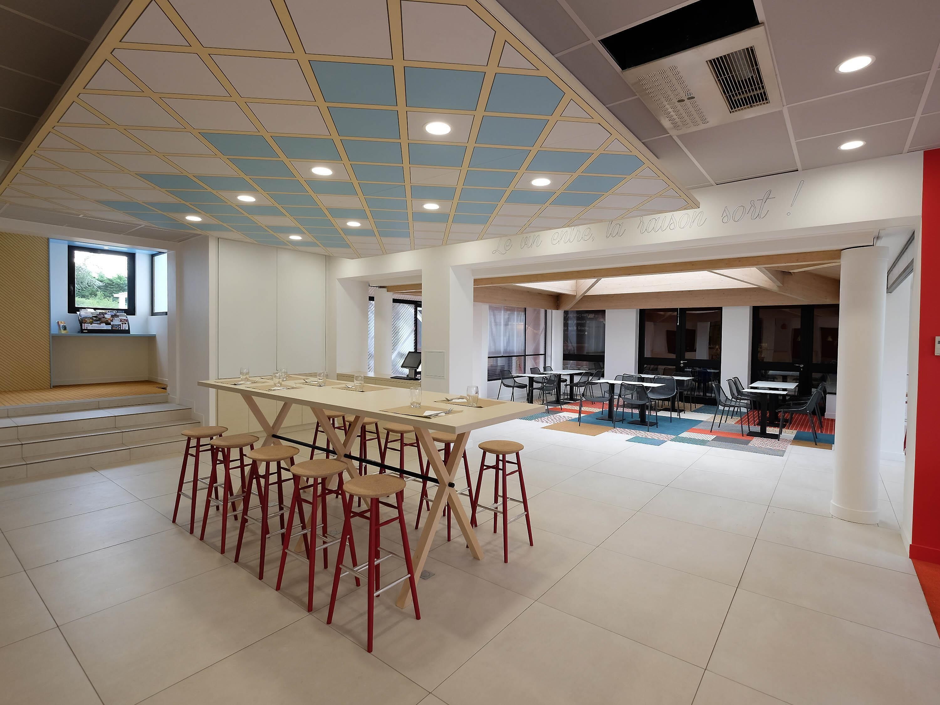 Een ruime kamer met een lange tafel en meerdere stoelen, een hoog plafond met geometrische patronen en een raam dat voor daglicht zorgt.