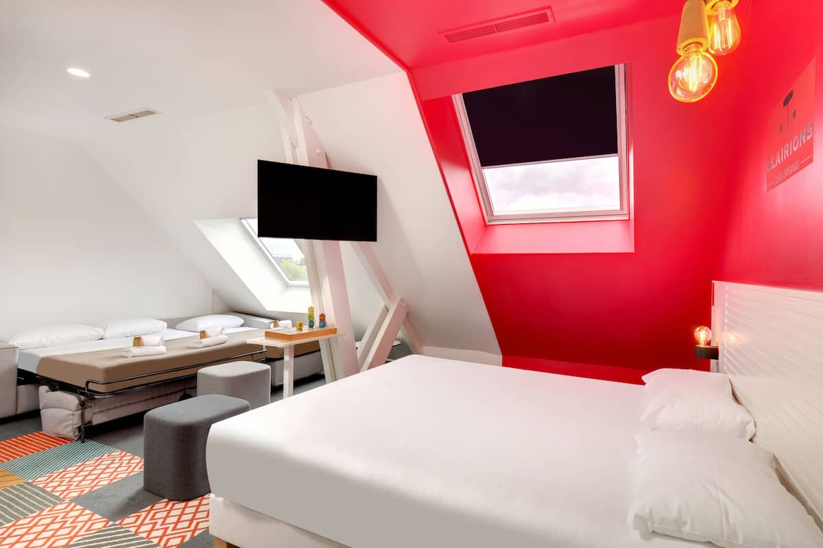 Ein modernes Hotelzimmer mit roter Akzentwand, einem großen Bett, einem Schreibtisch und einem an der Wand befestigten Fernseher.