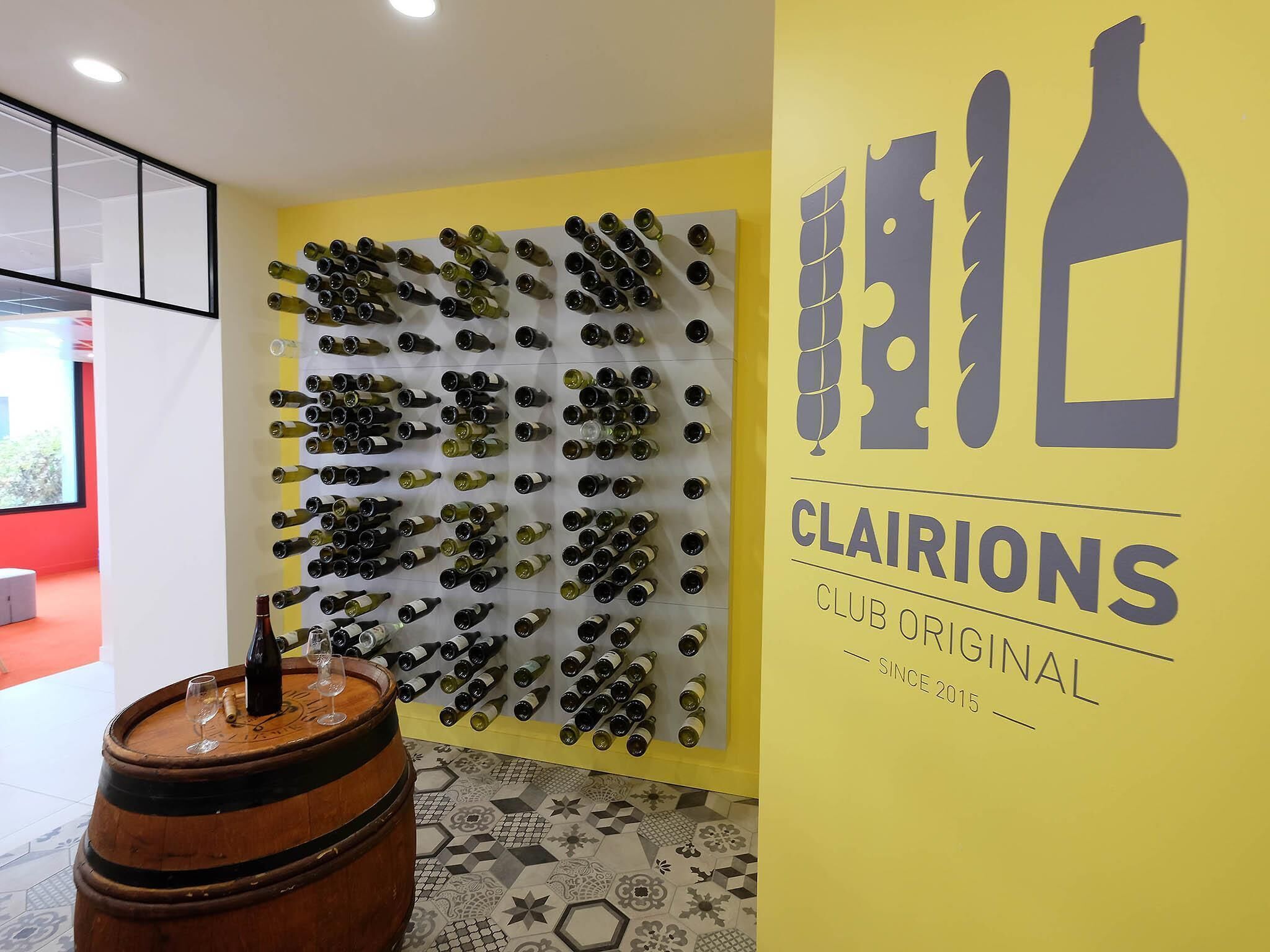 A wall with wine bottles arranged in rows, a wooden barrel with a wine glass, and a yellow wall with the text 'CLAIRIONS CLUB ORIGINAL SINCE 2015'.