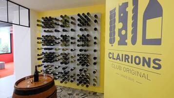 A wall with wine bottles arranged in rows, a wooden barrel with a wine glass, and a yellow wall with the text 'CLAIRIONS CLUB ORIGINAL SINCE 2015'.