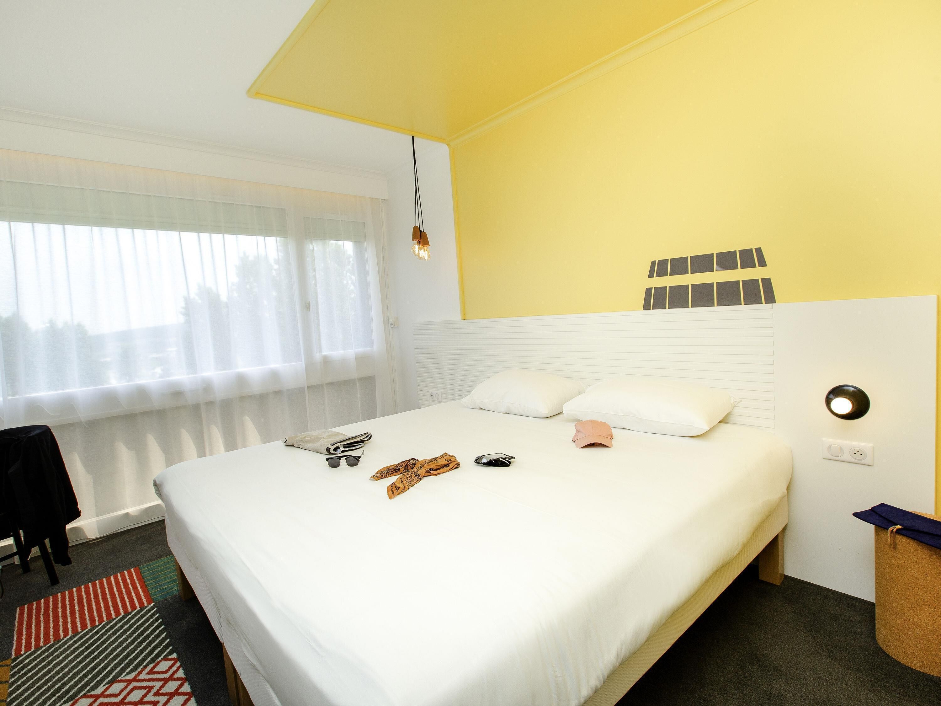 Una habitación de hotel con cama, ventana con cortinas translúcidas, mesita de noche y lámpara de pared.