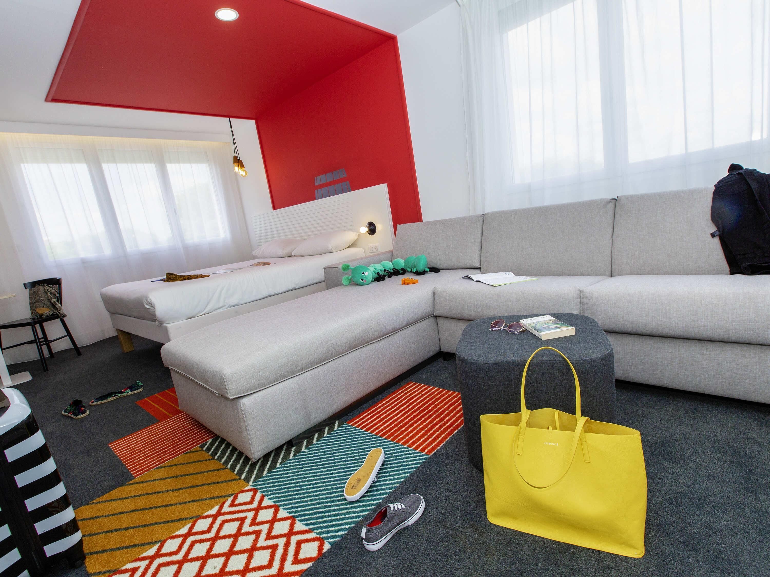 Un salon moderne avec un canapé gris, un mur d’accent rouge et un sac jaune posé sur le sol.