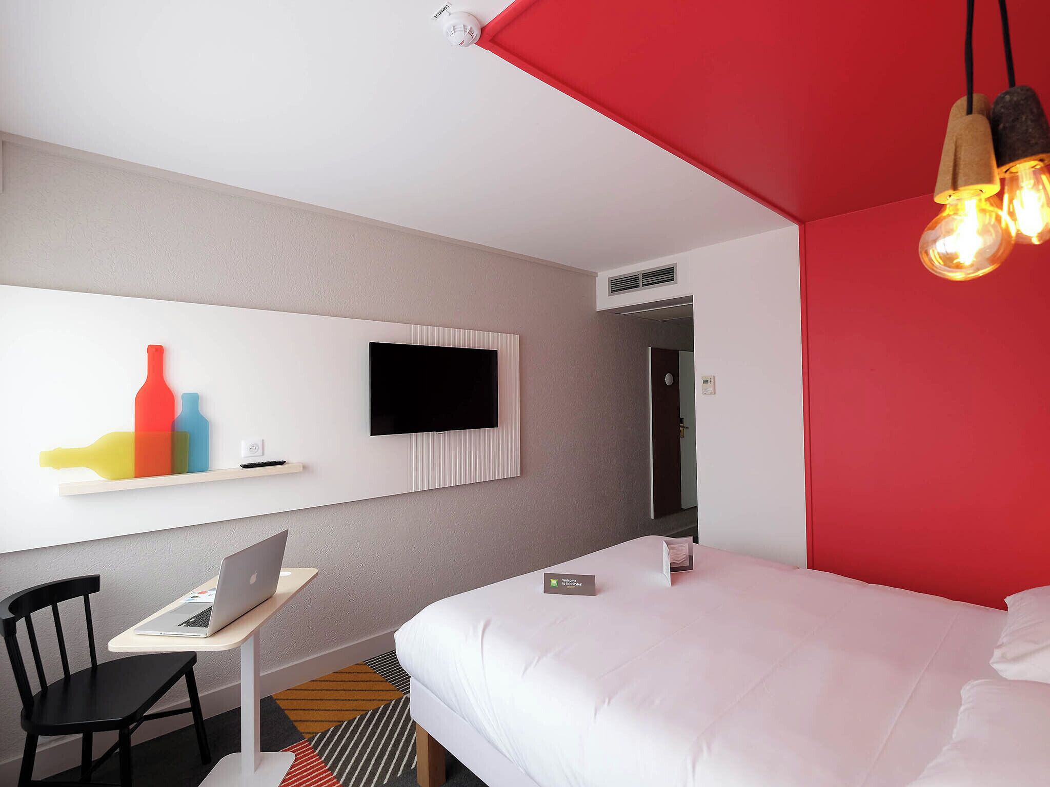 A hotel room with a bed, a desk with a laptop, a TV mounted on the wall, and a red accent wall.