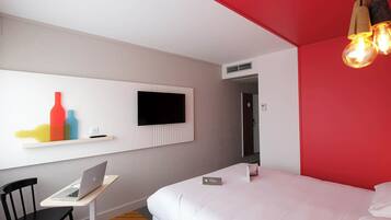 A hotel room with a bed, a desk with a laptop, a TV mounted on the wall, and a red accent wall.