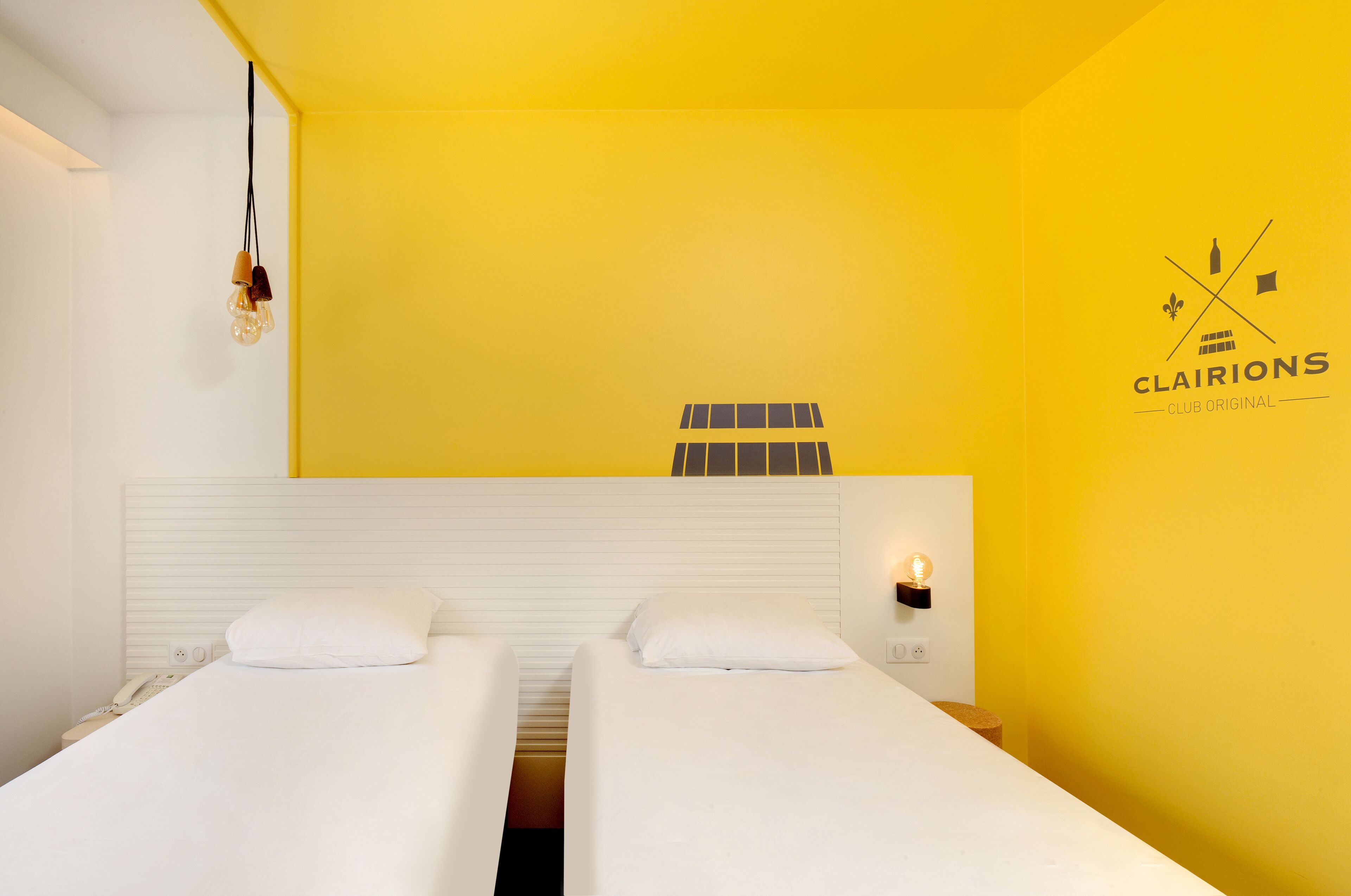 A hotel room with two beds, a white headboard, a yellow wall, and a logo on the wall.