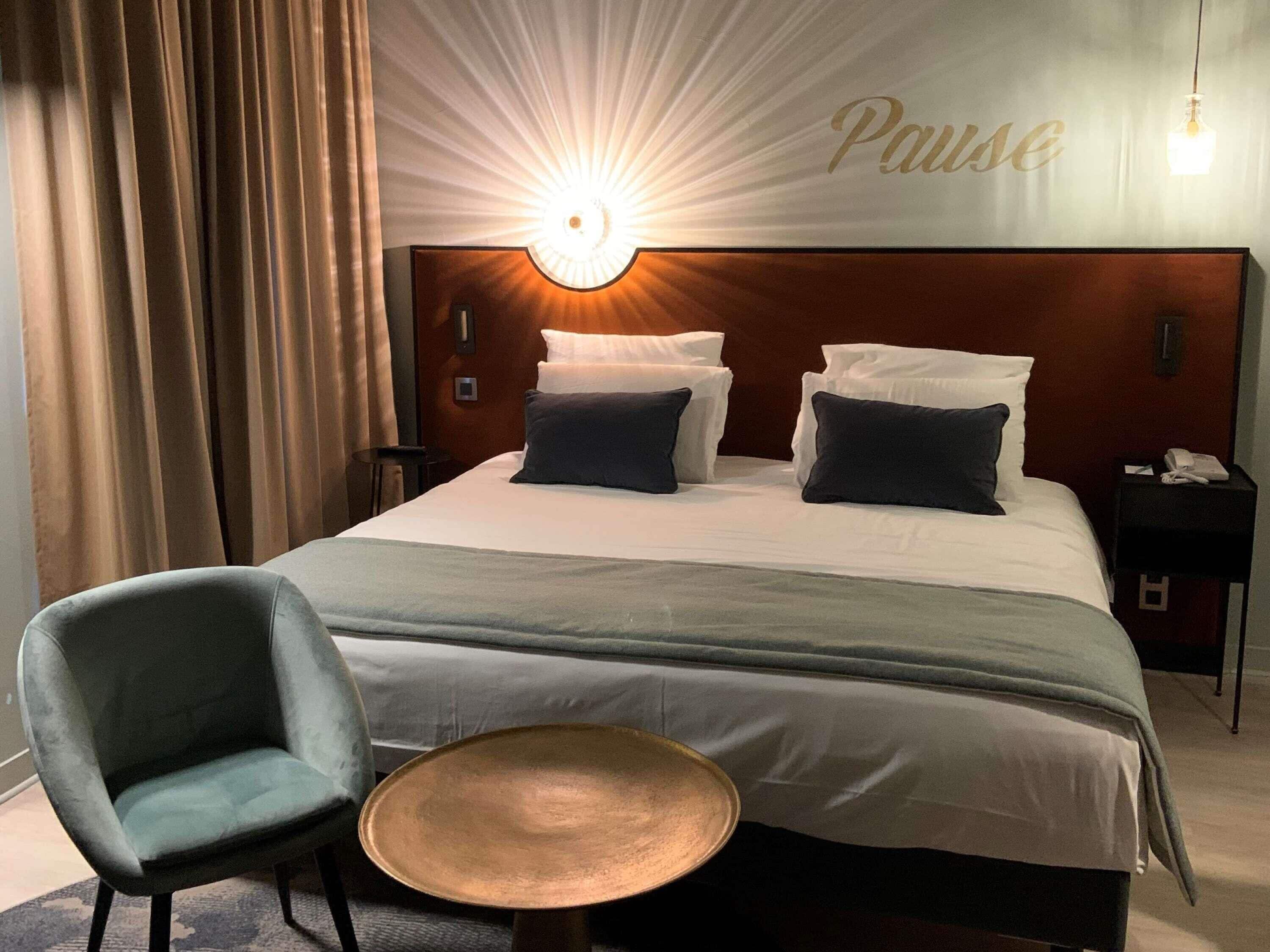 A hotel room with a bed, a chair, a small table, and a wall-mounted light.