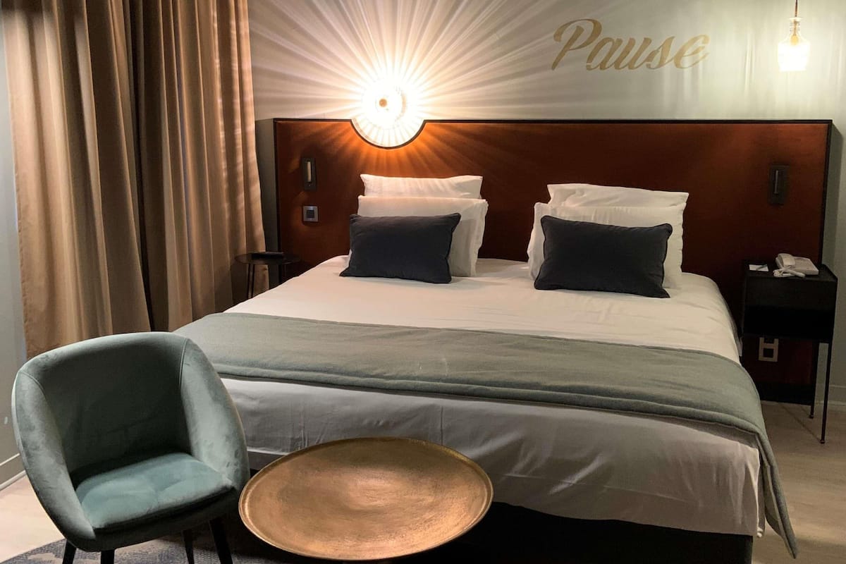 A hotel room with a bed, a chair, a small table, and a wall-mounted light.