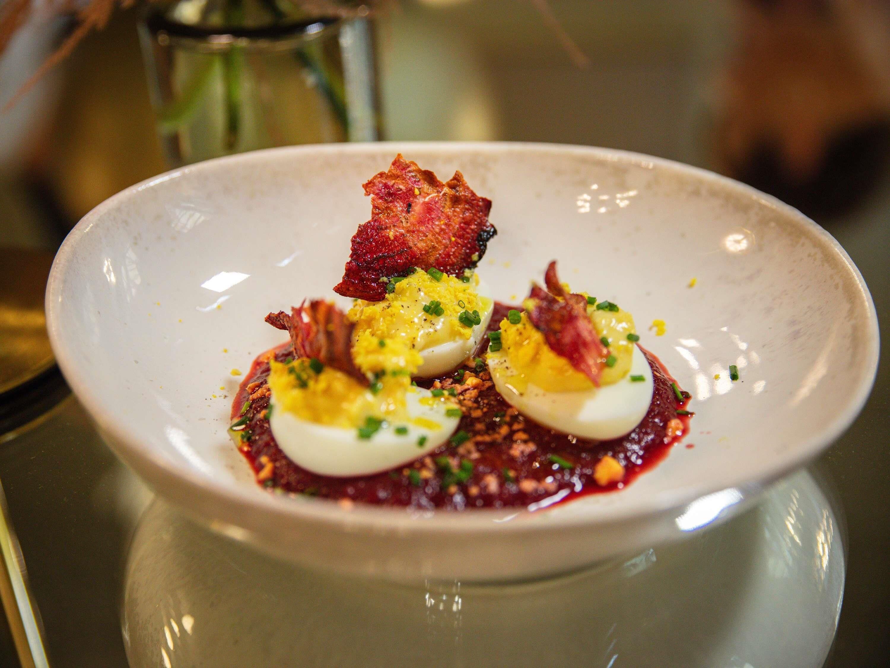 A white plate with three halves of a hard-boiled egg, garnished with chives and bacon, on a dark surface.
