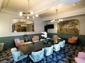 Un salón de conferencias con una mesa de mármol, sillas tapizadas, un espejo grande, una televisión y un mural en la pared.
