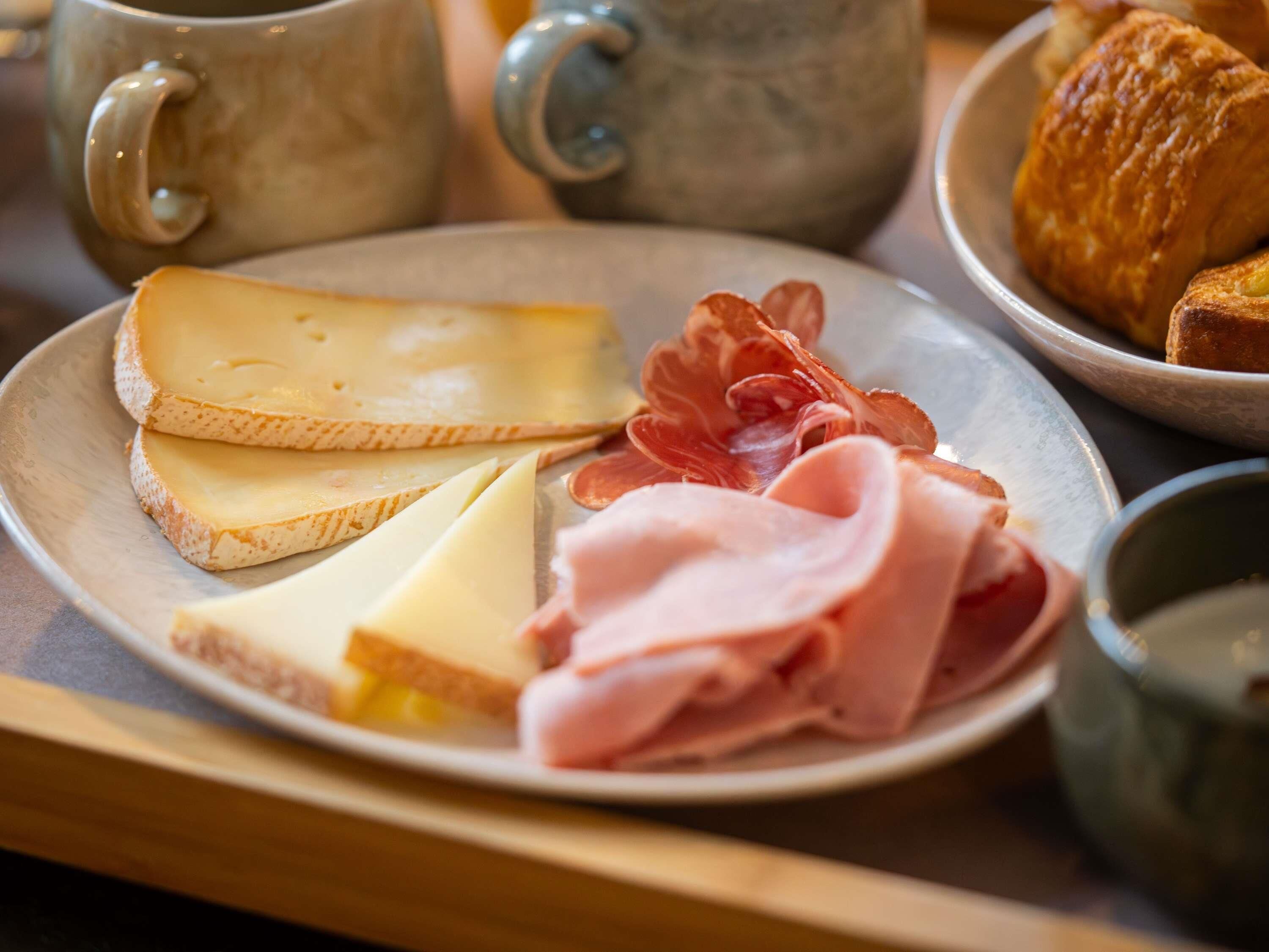 A plate with cheese, ham, and prosciutto, a croissant, and a mug on a table.