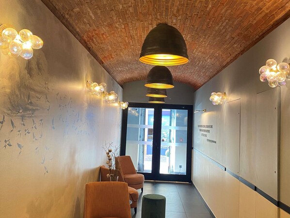 A hallway with brick ceiling, wall-mounted lights, a door with glass panels, and a seating area with chairs and a vase.