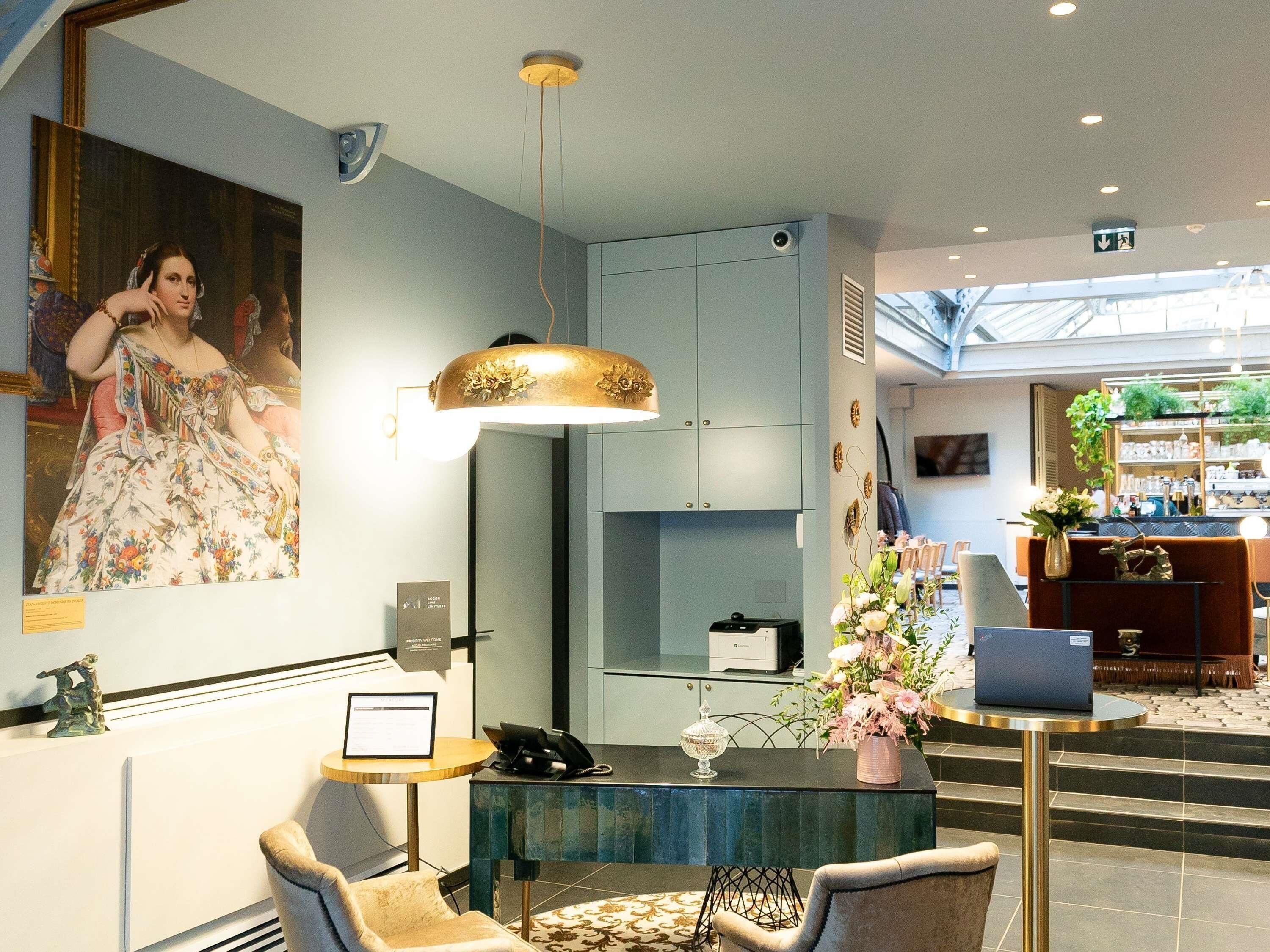 Un hall d’hôtel comprenant un comptoir d’accueil, un grand tableau accroché au mur, un lustre moderne et un escalier menant à l’étage supérieur.