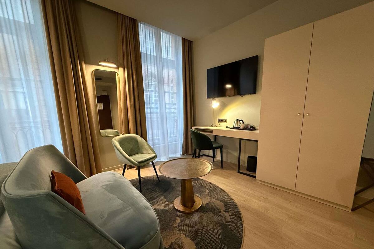 Ein modernes Hotelzimmer mit einer Couch, einem runden Couchtisch, einem Schreibtisch mit Stuhl, einem Flachbildfernseher und einem Kleiderschrank.