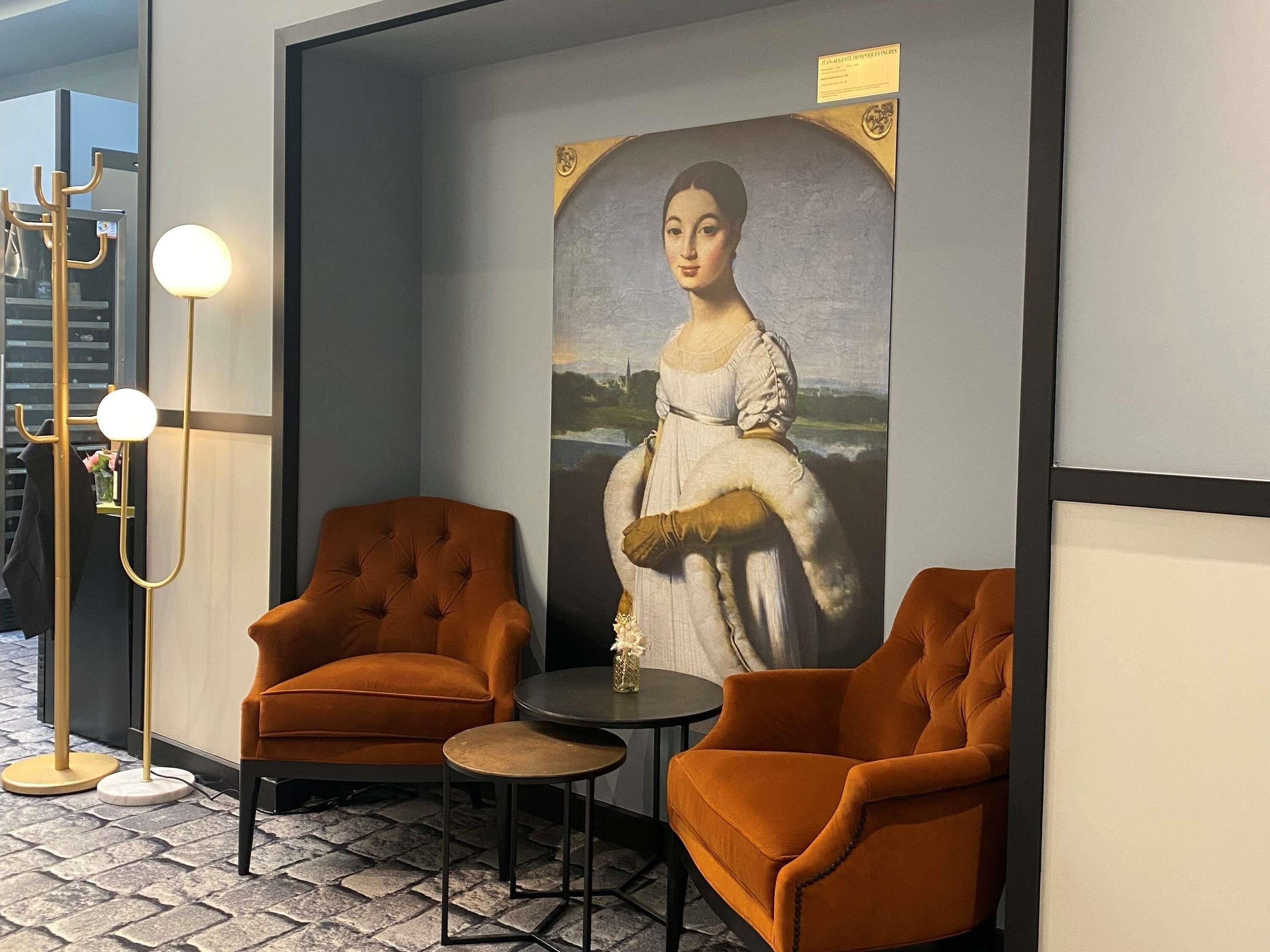 A room with two orange armchairs, a small round table, a floor lamp, and a large framed painting on the wall.
