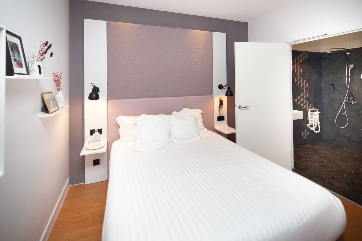 A modern bedroom with a large bed, bedside tables, a nightstand, a wall-mounted lamp, and a shower area with a tiled wall.