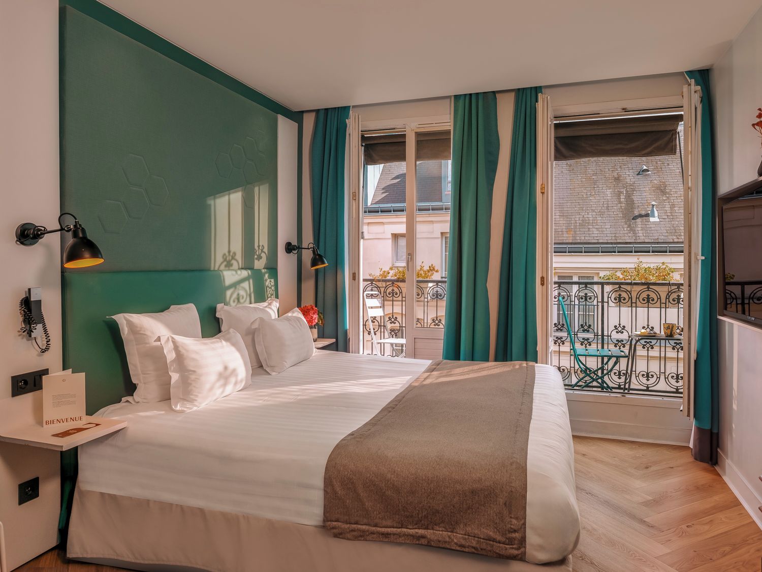Habitación de hotel con cama, mesita de noche, lámpara, ventana con cortinas y vista al balcón exterior.