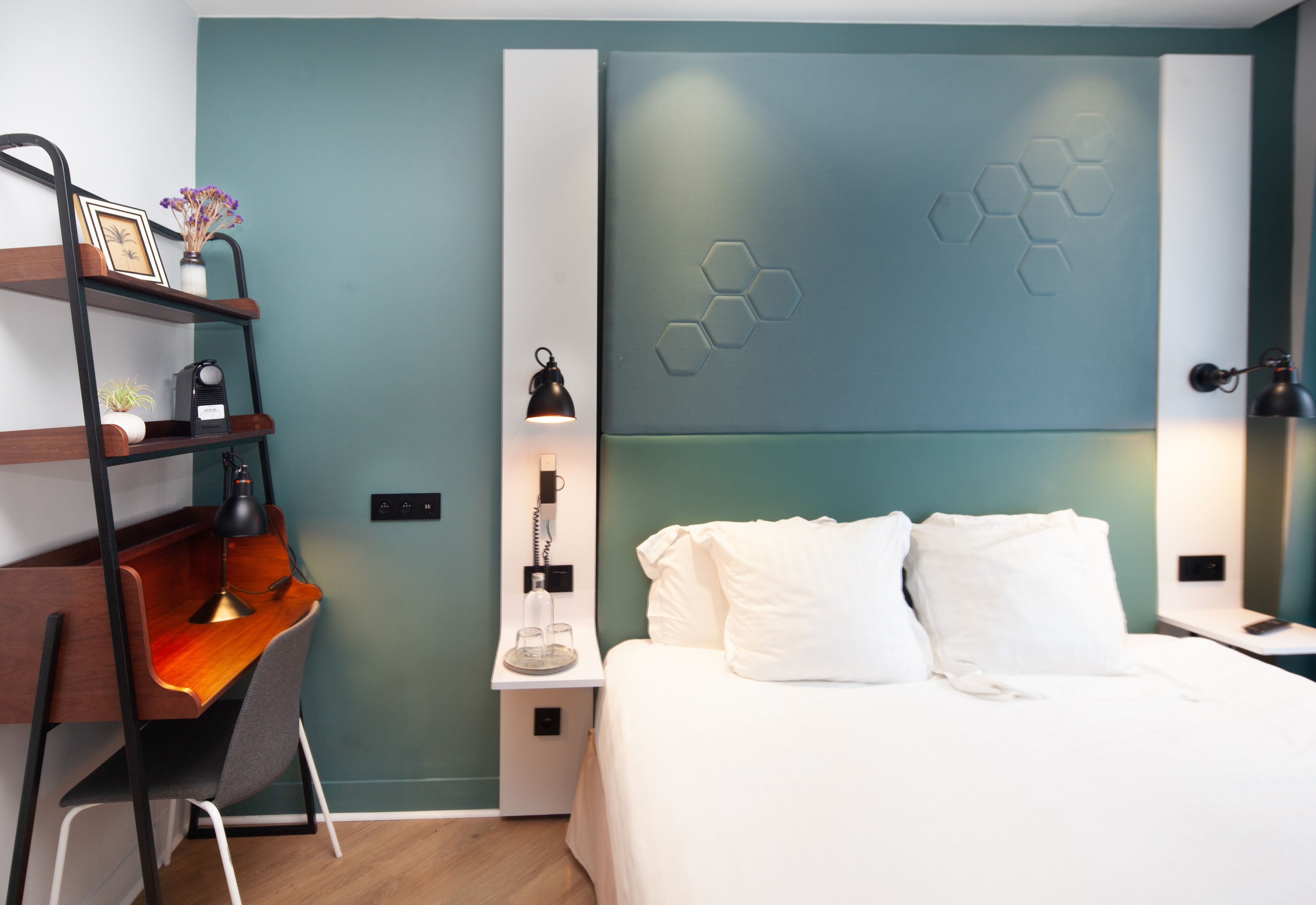 Una habitación de hotel moderna con una cama, un escritorio, una silla, una mesita de noche y una pared con diseños hexagonales.