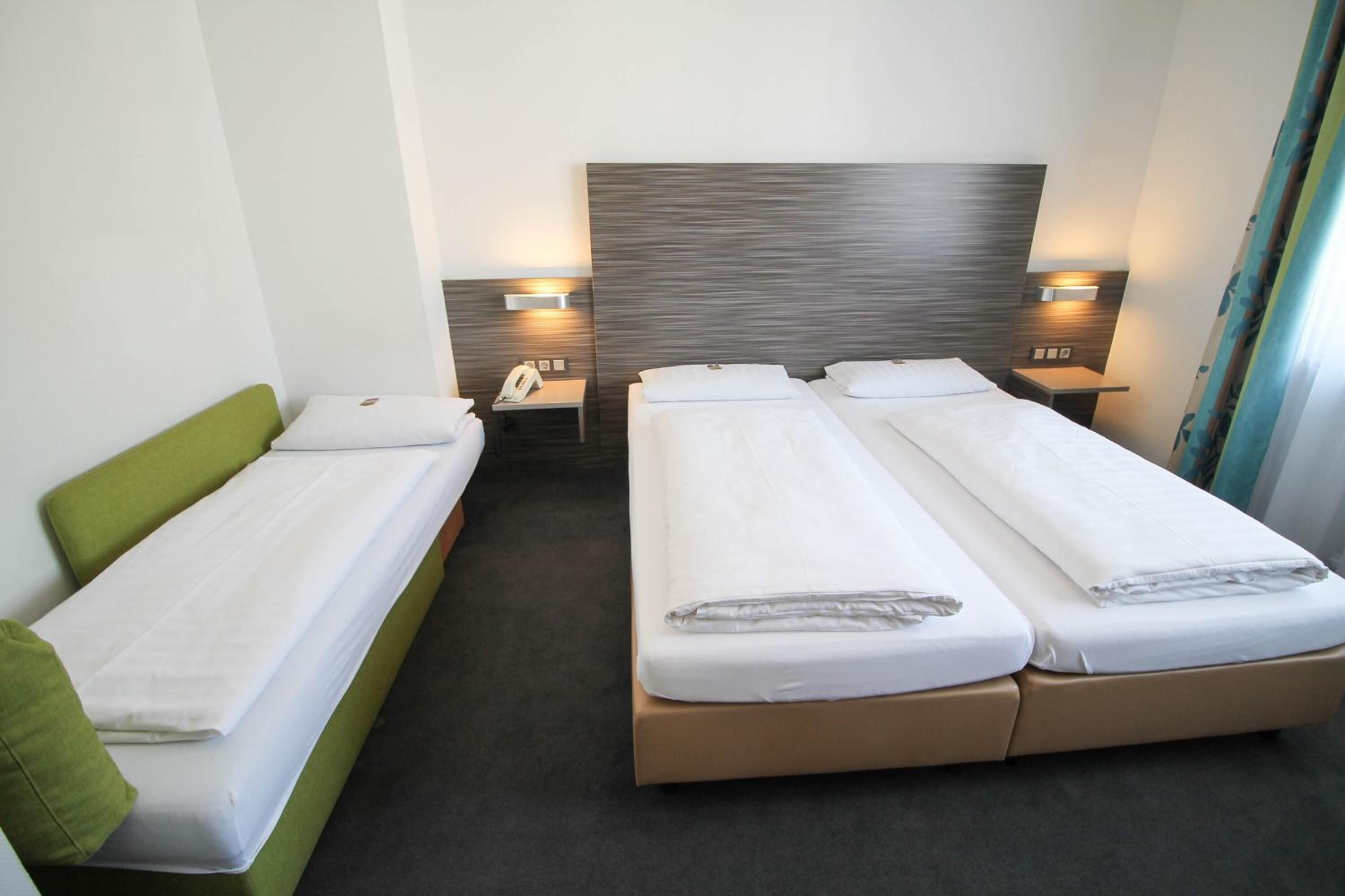 Habitación de hotel con dos camas individuales, un sofá verde, un cabecero de madera, lámparas de noche y un teléfono en la mesita de noche.